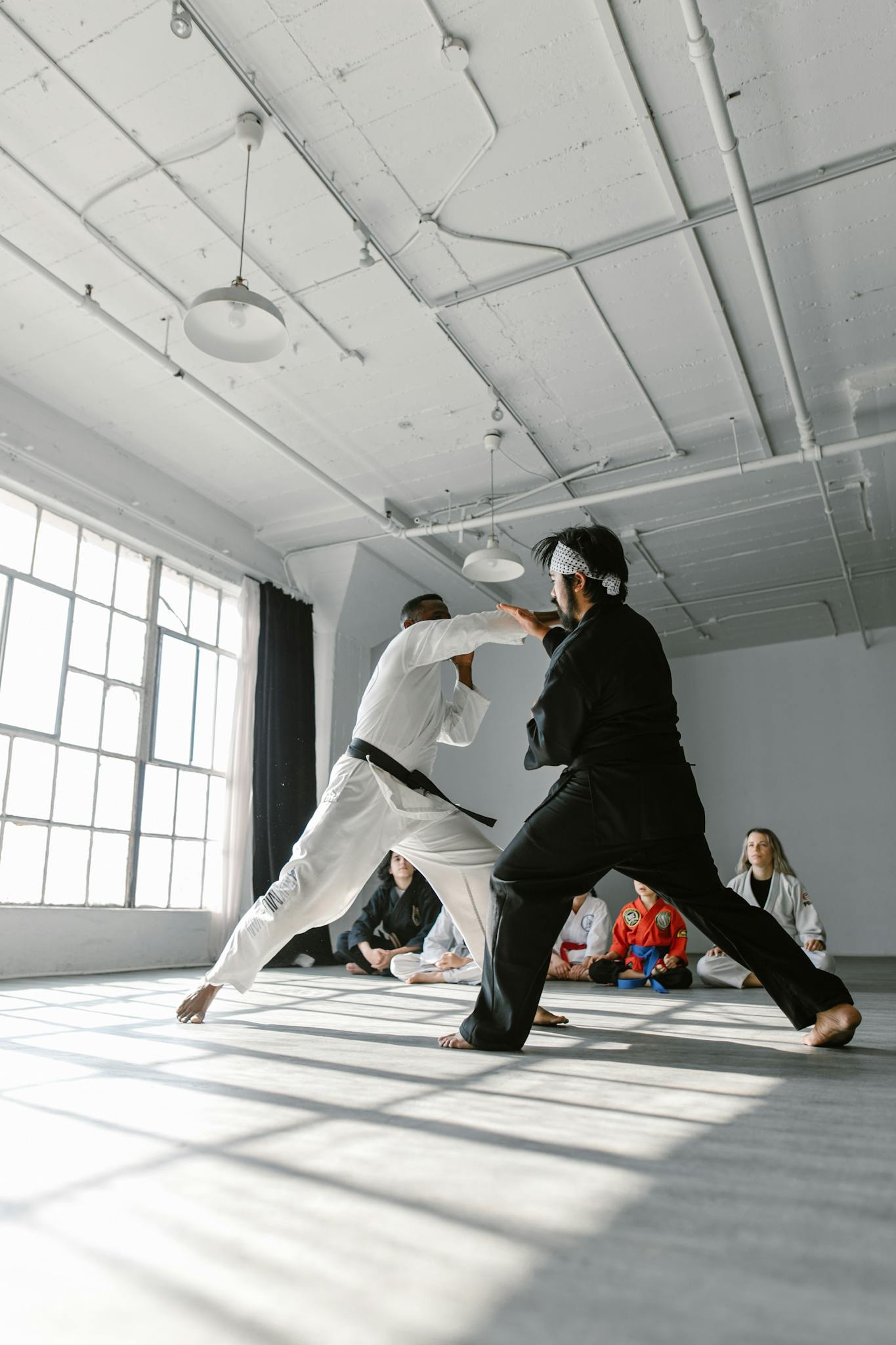 Martial artists practicing sparring in a sunlit gym. White and black uniforms add contrast.