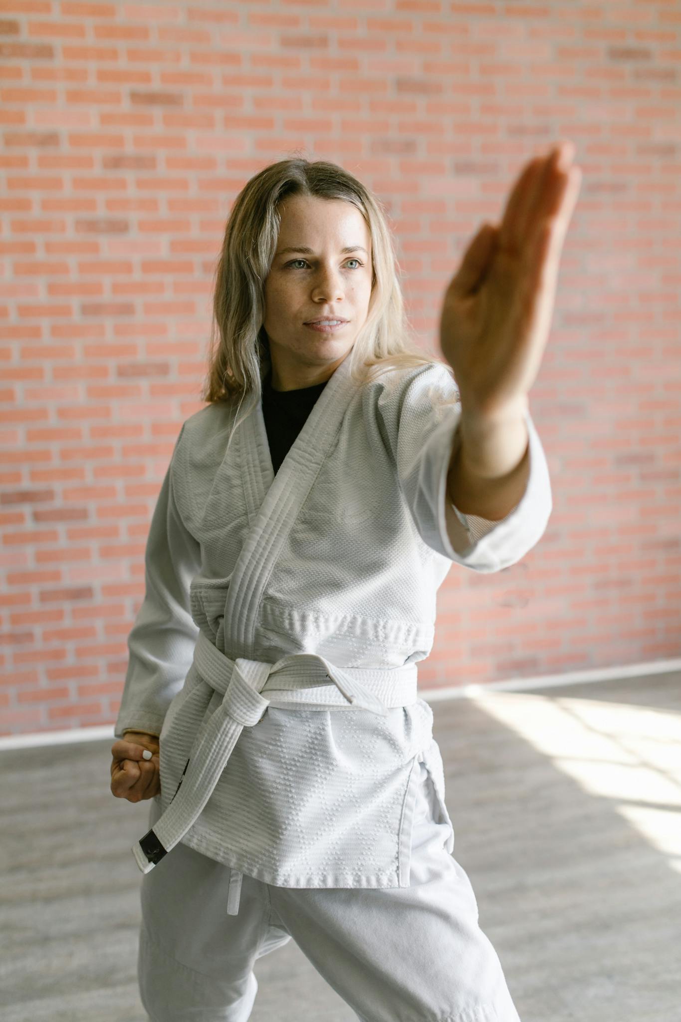 Woman practicing martial arts against a brick wall, in a white uniform and belt.