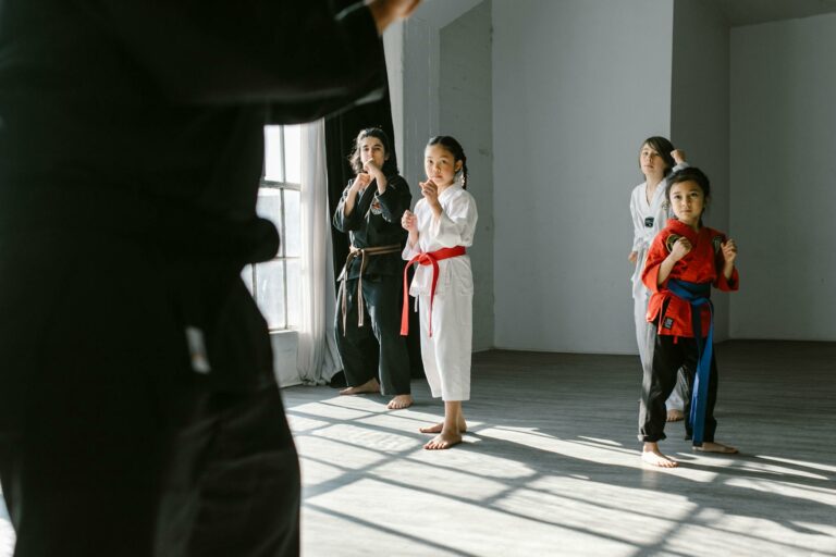 Group of children practicing martial arts indoors, guided by an instructor, in a sunlight-lit dojo.